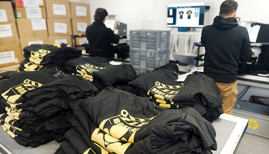 lot de tee-shirts imprimés posés sur un plan de travail, à l'arrière plan les salariés impriment les textiles pour un évènement 